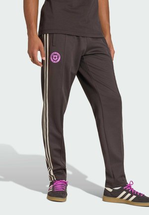 Persona che indossa pantaloni della tuta Adidas neri con strisce beige, un emblema circolare rosa sulla coscia e sneakers nere con lacci viola.