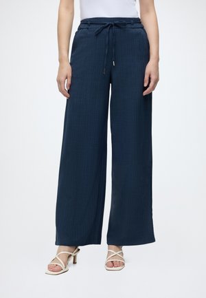 Femme portant un pantalon large bleu marine avec une taille à cordon et des sandales blanches à talons, debout devant un fond blanc.