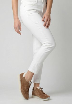 Osoba ubrana w białe, dopasowane spodnie i brązowe zamszowe trampki, stojąca z jedną nogą lekko uniesioną na jednolitym szarym tle.