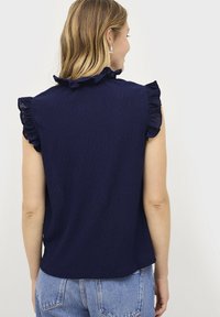 Marineblauw mouwloos topje met gerimpelde schouderaccenten, textuurstof en een ontspannen pasvorm, gecombineerd met lichtblauwe high-waisted jeans.