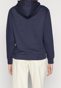 Felpa navy con tasca a marsupio e cappuccio con cordino, realizzata in morbido tessuto misto cotone. Polsini e orlo a coste. Indossata con pantaloni chiari.