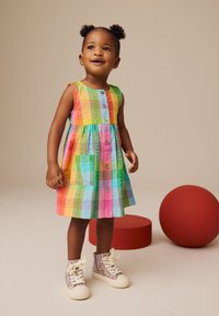 Kolorowa, paskowana sukienka bez rękawów w odcieniach zieleni, różu, niebieskiego i żółtego, z guzikami z przodu i bocznymi kieszeniami. Noszona z błyszczącymi trampkami.