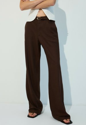 Personne portant un pantalon large marron taille haute, une ceinture noire, un haut blanc et des sandales noires, debout les bras croisés.