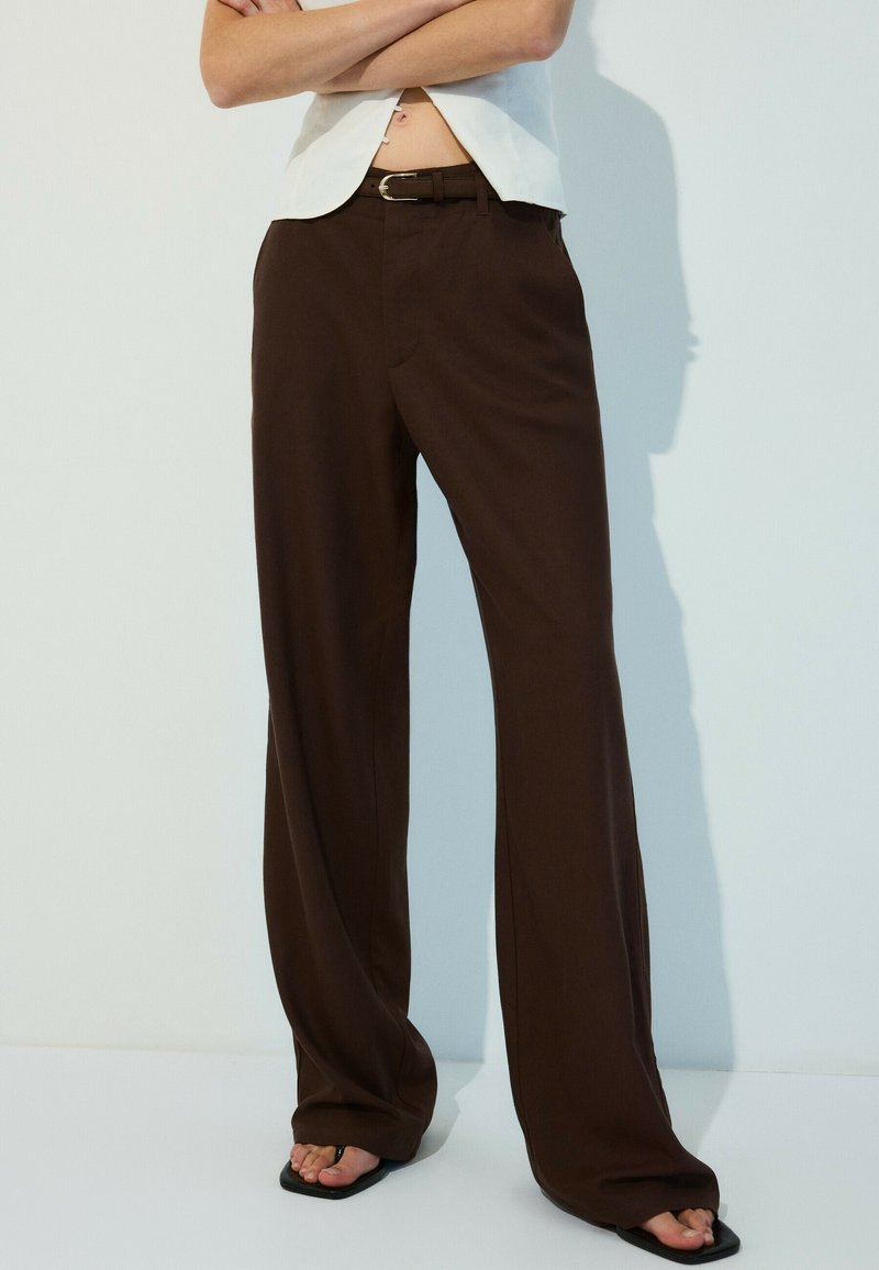 Personne portant un pantalon large marron taille haute, une ceinture noire, un haut blanc et des sandales noires, debout les bras croisés.