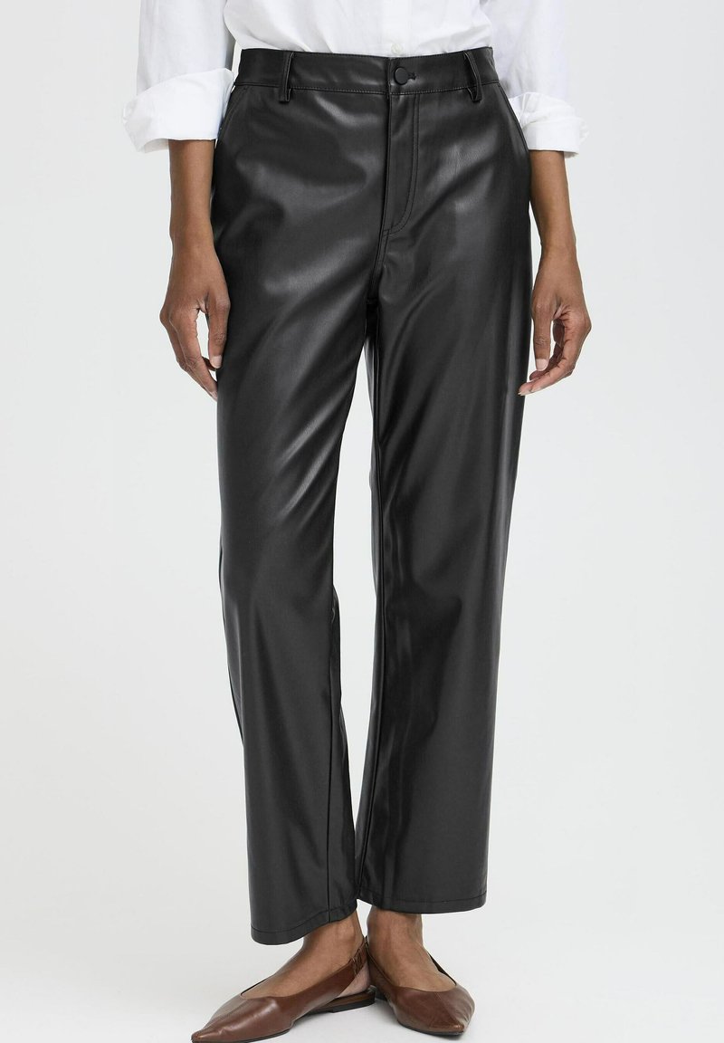 Personne portant un pantalon droit en cuir noir, une chemise blanche aux manches retroussées et des chaussures plates marron, debout sur un fond clair.