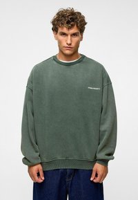 Sweatshirt vert surdimensionné en tissu doux, avec un col ras du cou, des épaules tombantes et un petit logo brodé blanc sur le devant.