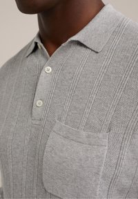 Detailní záběr osoby nosící světle šedé pletené polo tričko s texturou, bílými knoflíky a kapsou na prsou.