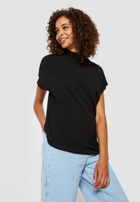 Haut noir à manches courtes avec un col montant, fabriqué à partir d'un tissu léger. Modèle portant un jean bleu clair, les mains dans les poches, cheveux bouclés visibles.