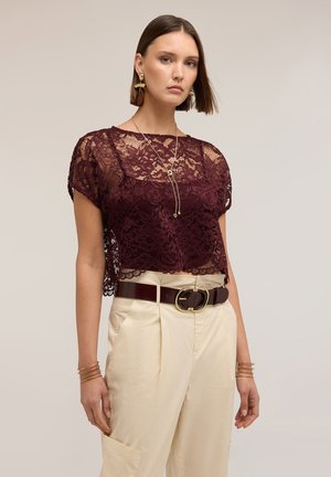 Nainen, joka pukeutunut läpikuultavaan viininpunaiseen pitsitoppiin, kermaan sävyisiin korkeavyötäröisiin housuihin, joissa tumma vyö, kultaiset korvakorut, kaulakoru ja rannekorut, poseeraa yksiväristä taustaa vasten.