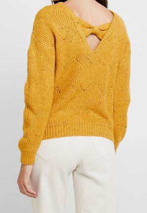 Pull en maille jaune moutarde avec un design en V au dos, orné d'un nœud, de poignets côtelés et d'un motif texturé. Porté sur un jean blanc.
