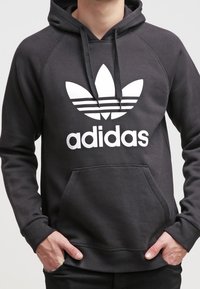 Svart huvtröja med en framficka, med en stor vit Adidas-logotyp och tre ränder ovanför logotypen. Tillverkad av mjukt material, ribbade muddars.