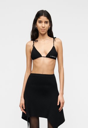 Mujer con cabello largo y oscuro, vistiendo un top de bikini negro con forma triangular y texto, junto con una falda negra asimétrica acanalada, sobre un fondo liso.