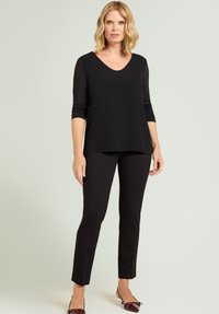 Donna che indossa un top nero a maniche lunghe e pantaloni neri aderenti, in piedi con un'espressione neutra su uno sfondo chiaro e semplice.