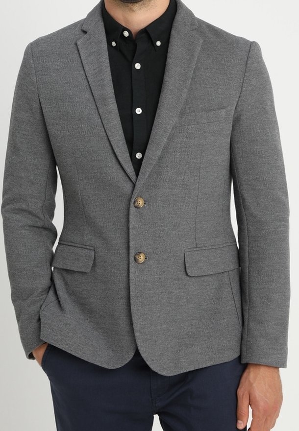 Blazer gris avec un tissu texturé, deux poches avant et des boutons marron. Porté par-dessus une chemise noire à col. Coupe ajustée.