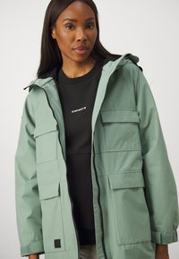 Manteau vert avec capuche, plusieurs poches avant et fermeture éclair. Porté par-dessus un haut noir à côtes. Tissu texturé avec une finition lisse.