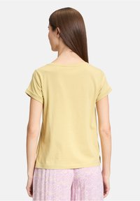 Gul bomulds t-shirt med korte, rullede ærmer og rund halsudskæring, vist fra ryggen, sat sammen med mønstrede lavendelbukser.