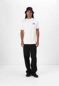 White cotton t-shirt featuring a colorful graphic on the chest, paired with black wide-leg trousers and a black bucket hat. Simple design.