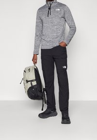Grauer Pullover mit langen Ärmeln und Reißverschluss, schwarze Cargohose und ein zweifarbiges Rucksack. Die Hose hat Seitentaschen; die Schuhe sind schwarze Sportschuhe.