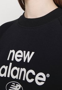 Musta huppari, jossa on pyöreä kaula-aukko ja raglan-hihat. Näyttää "new balance" -logon paksuilla valkoisilla kirjaimilla. Pehmeä kankaan pinta.