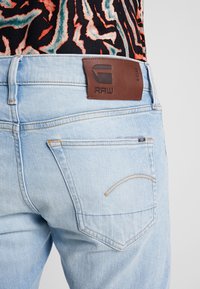 Světle modré džínové kalhoty z denimového materiálu s koženou etiketou RAW, kontrastním šitím, pětipocketovým designem a viditelnými detaily zadních kapes.