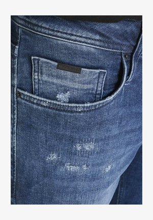 Dunkelblaue Jeans aus Denim mit einer distressierten Textur, ausgestattet mit einer kleinen Tasche mit einem schwarzen Etikett und sichtbaren Nähten am Bund.