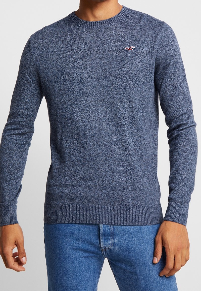 Pull en tricot bleu avec un col rond et un ourlet côtelé. Présente un petit logo sur la poitrine et des manches longues. Texture lisse, sans motifs.