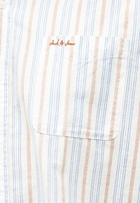 Λευκό πουκάμισο με κάθετες μπλε και πορτοκαλί ρίγες, που διαθέτει μια τσέπη στο στήθος με πορτοκαλί κεντημένο κείμενο από πάνω της.