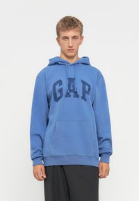 Un joven con cabello rubio corto, vestido con una sudadera azul de GAP y pantalones oscuros, de pie frente a un fondo blanco liso.