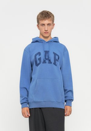 Giovane uomo con capelli biondi corti, che indossa un felpa blu GAP e pantaloni scuri, in piedi di fronte a uno sfondo bianco semplice.