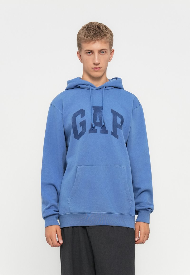 Un joven con cabello rubio corto, vestido con una sudadera azul de GAP y pantalones oscuros, de pie frente a un fondo blanco liso.