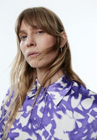 Paarse en witte blouse met patroon en kraag, met een satijnen textuur en knoopsluiting. Het haar bedekt gedeeltelijk het gezicht van het model.