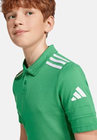 Grünes Poloshirt mit weißen Streifen auf den Schultern. Verfügt über einen Kragen, eine Zwei-Knopf-Leiste und ein markiertes Detail am Ärmel.
