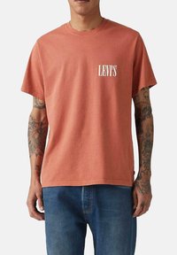 Camiseta de algodón color óxido con un corte relajado, mangas cortas y un logotipo blanco "LEVI'S" en el lado izquierdo del pecho. Combinada con vaqueros azules.