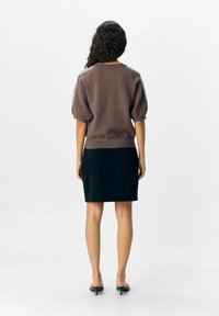 Un pull en tricot marron avec des manches courtes bouffantes et des embellissements contrastants, associé à une jupe noire ajustée, porté avec des sandales noires élégantes.