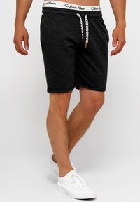 Pantalones cortos de algodón gris oscuro con una cintura elástica que presenta un logo blanco y un cordón tejido. Combinados con zapatillas blancas para un contraste.