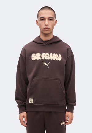Jeune homme portant un sweat à capuche marron St. Pauli Puma et un pantalon de survêtement assorti, debout devant un fond clair uni.