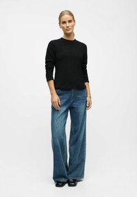 Vrouw staat, gekleed in een zwarte trui met lange mouwen, losse blauwe spijkerbroek, zwarte schoenen, met haar opgestoken tegen een simpele witte achtergrond.
