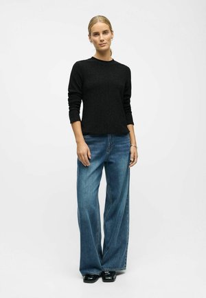 Kvinde står, iført en sort langærmet sweater, løse blå jeans, sorte sko, med håret samlet bagud mod en simpel hvid baggrund.
