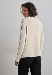 Vrouw met kort, krullend haar, gekleed in een crèmekleurige, geribbelde trui met donkere streepdetails op de mouwen, zwarte broek, staande tegen een effen grijze achtergrond.