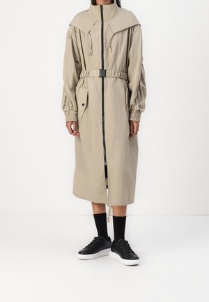 Osoba oblečená v béžovém dlouhém kabátu s páskem, předním zipem a kapsou s klopou, doplněném černými ponožkami a černými teniskami s bílými podrážkami.