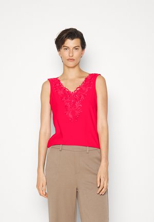 Top sem mangas vermelho com decote em V e detalhes florais bordados. Tecido liso e leve complementado por calças beges.