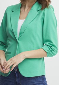 Zelený, lehký blazer s přiléhavým střihem, s klopami a rukávy do tří čtvrtin. Obsahuje jeden knoflík a přední kapsu.