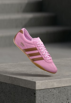 Ροζ αθλητικό παπούτσι Adidas Tokyo με καφέ ρίγες και σόλα, αιωρούμενο πάνω από ένα τσιμεντένιο χείλος σε θολό γκρι φόντο.