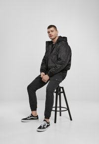 Giacca nera imbottita con cappuccio, pantaloni neri a sigaretta e sneakers nere con dettagli bianchi, seduto su uno sgabello nero contro uno sfondo chiaro.