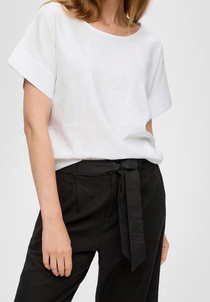 Nainen, jolla on valkoinen lyhythihainen paita ja korkeavyötäröiset mustat housut, joissa on sidottu kangasvyö, kuvattu kaulasta lantioihin.