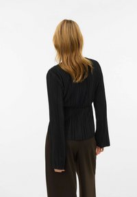 Svart plissért langermet topp med innsnevret midje, laget av teksturert stoff og med flagrende ermer, kombinert med brune vide bukser.