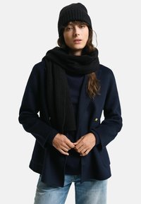 Cappotto navy doppiopetto, sciarpa nera a coste e berretto, abbinati a un maglione scuro e a jeans azzurri chiari. Tessuti morbidi e design strutturato.