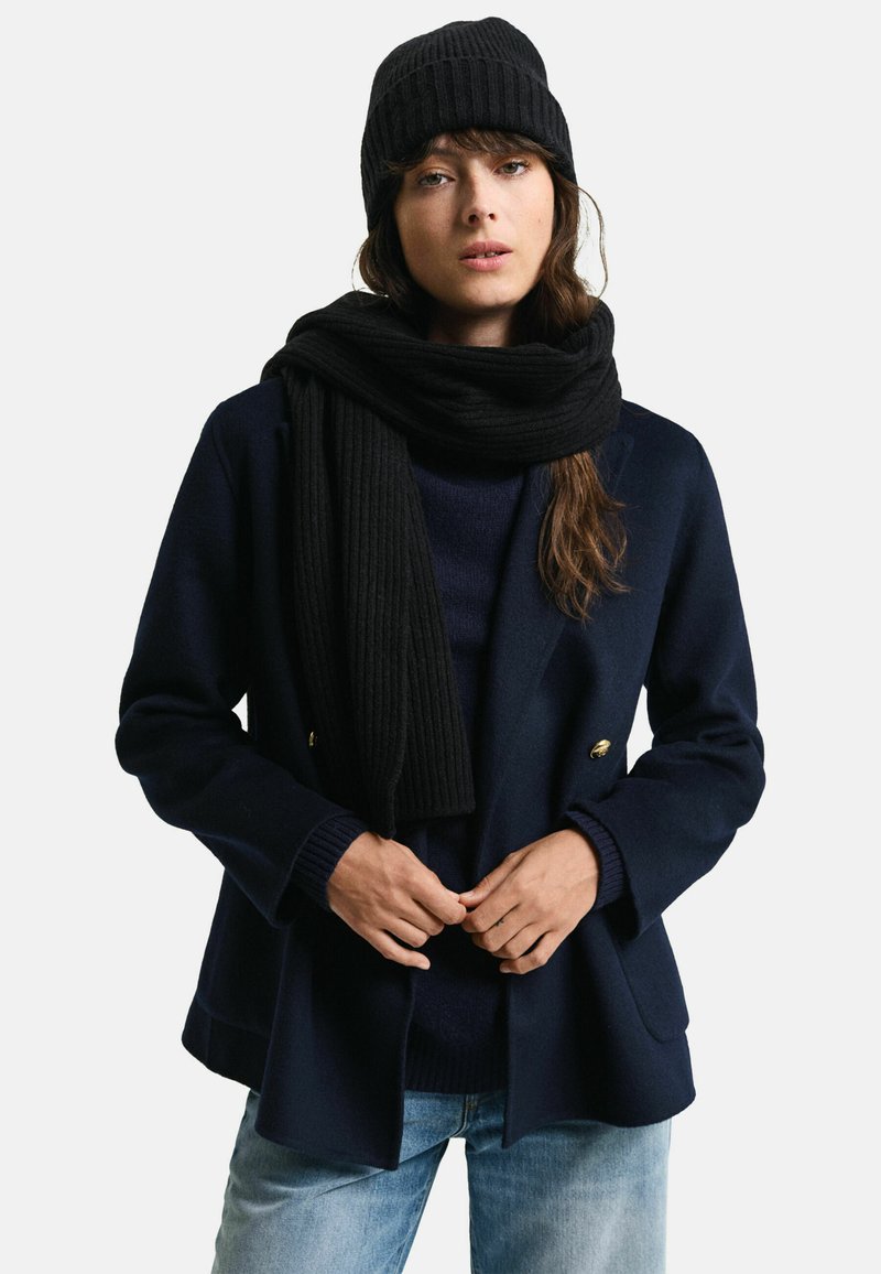 Cappotto navy doppiopetto, sciarpa nera a coste e berretto, abbinati a un maglione scuro e a jeans azzurri chiari. Tessuti morbidi e design strutturato.