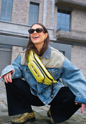 Vrouw met een zonnebril, een tweekleurige spijkerjas, gele crossbodytas, zwarte wijde broek en geblokte laarzen hurkt buiten bij stenen gebouwen.