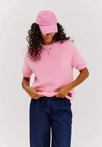 Ružový svetr krátkych rukávov vyrobený z mäkkého materiálu s okrúhlym výstrihom, skombinovaný s tmavomodrými nohavicami s vysokým pásom a zodpovedajúcou ružovou čiapkou.